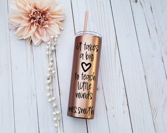 Puede incluir: Un vaso de acero inoxidable en color oro rosa con tapa transparente y pajita a juego. El vaso presenta un texto en negro que dice "it takes a big heart to teach little minds" seguido del nombre "ms. smith" en letra cursiva.