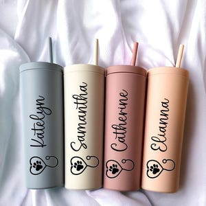 May include: Four matte finish tumblers with straws in muted blue, cream, dusty rose, and peach. Each tumbler features a black script name and a stethoscope icon shaped like a heart with a paw print inside. The names are Katelyn, Samantha, Catherine, and Elianna.