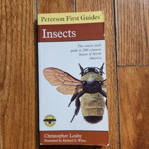 May include: A paperback book titled "Insects" from Peterson First Guides. The cover features a detailed illustration of a bee with its wings spread. The book is a concise field guide to 200 common insects of North America.