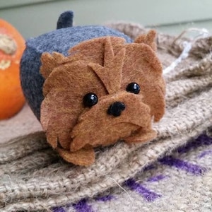 May include: A brown and gray felt toy dog with black eyes and a black nose. The dog is sitting on a burlap surface.