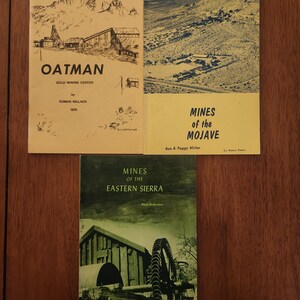 May include: Three softcover books with historical themes. The top left book is yellow with a black line drawing of a mountain and the text OATMAN GOLD MINING CENTER. The top right is yellow with a landscape photo and MINES of the MOJAVE. The bottom is green with a photo of mining equipment and MINES OF THE EASTERN SIERRA.