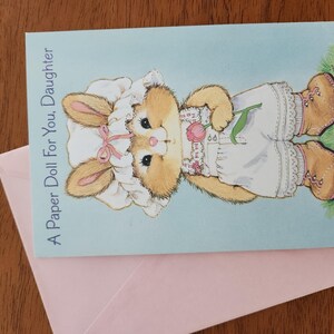 May include: A greeting card with a light blue background featuring a cartoon bunny wearing a bonnet and holding a tulip. The card reads "A Paper Doll For You, Daughter." A pink envelope is beneath the card.