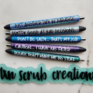 May include: Five glitter pens with different sayings on them. The sayings are: "Just another day in paradise", "Pretty good at bad decisions", "Don't be salty... that's my job", "Caution... I have no filter", and "Please don't make me do stuff". The pens are arranged vertically, with the first pen at the top and the last pen at the bottom. The pens are all black with glitter accents. The background is a light gray surface. The text "ton scrub creations" is written in black on a light blue background.