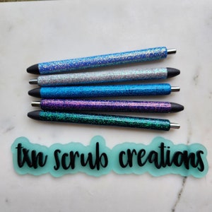 May include: Five glitter pens in various shades of blue, purple, and green. The pens have black caps and are arranged in a row on a white surface. The text "txn scrub creations" is visible in the lower part of the image.