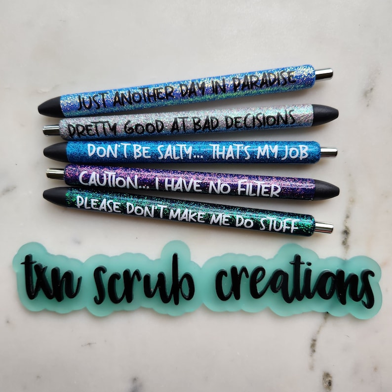 May include: Five glitter pens with different sayings on them. The sayings are: "Just another day in paradise", "Pretty good at bad decisions", "Don't be salty... that's my job", "Caution... I have no filter", and "Please don't make me do stuff". The pens are arranged vertically, with the first pen at the top and the last pen at the bottom. The pens are all black with glitter accents. The background is a light gray surface. The text "ton scrub creations" is written in black on a light blue background.