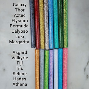 May include: A collection of 15 glitter-filled tubes, each with a different color and a name written below. The names include Galaxy, Thor, Aztec, Elysium, Bermuda, Calypso, Loki, Margarita, Asgard, Valkyrie, Fiji, Iris, Selene, Hades, and Athena.