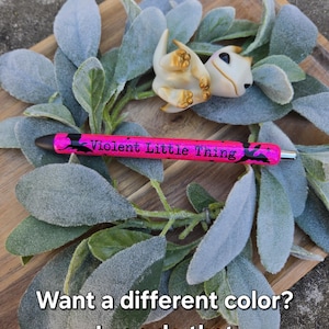 May include: A small white dragon figurine with gold accents, lying on a bed of green and gray leaves. The text "Violent Little Thing" is written on a pink pen.