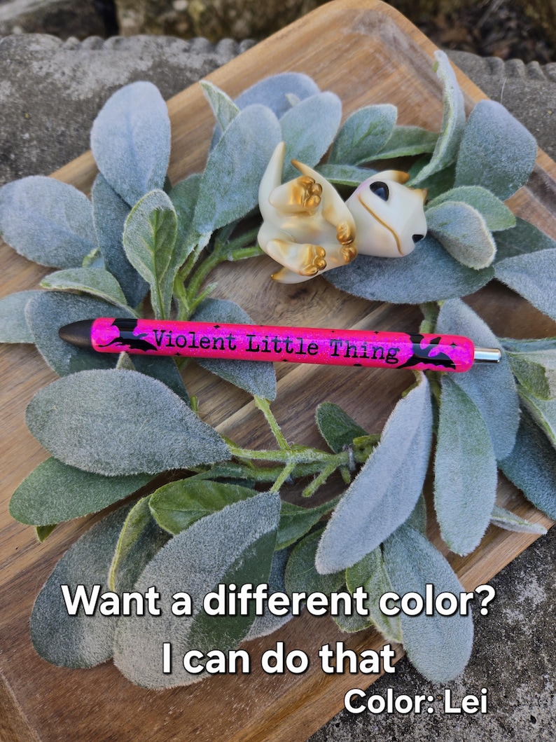 May include: A small white dragon figurine with gold accents, lying on a bed of green and gray leaves. The text "Violent Little Thing" is written on a pink pen.