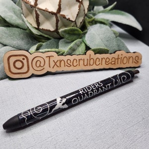 May include: A black pen with white glitter and the words "Riders Quadrant" printed on it. The pen is next to a wooden sign with the Instagram handle "@Txnscrubcreations" engraved on it.