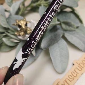 May include: A black and white pen with purple glitter and the text "Violent Little Thing" written in white on the side.