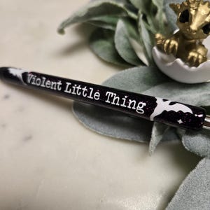 May include: Black pen with white glitter and a white bat design. The pen has the text "Violent Little Thing" written on it.