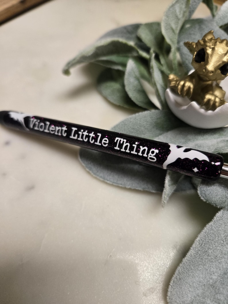 May include: Black pen with white glitter and a white bat design. The pen has the text "Violent Little Thing" written on it.