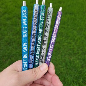 May include: Six glitter pens with different sayings on them. The sayings are: "Don't be salty... That's my job", "Just another day in paradise", "Please don't make me do stuff", "Decisions", "Pretty good at bad decisions", and "Caution... I have no filter".