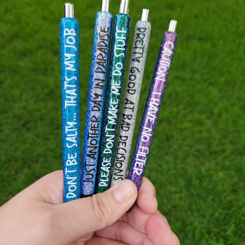 May include: Six glitter pens with different sayings on them. The sayings are: "Don't be salty... That's my job", "Just another day in paradise", "Please don't make me do stuff", "Decisions", "Pretty good at bad decisions", and "Caution... I have no filter".