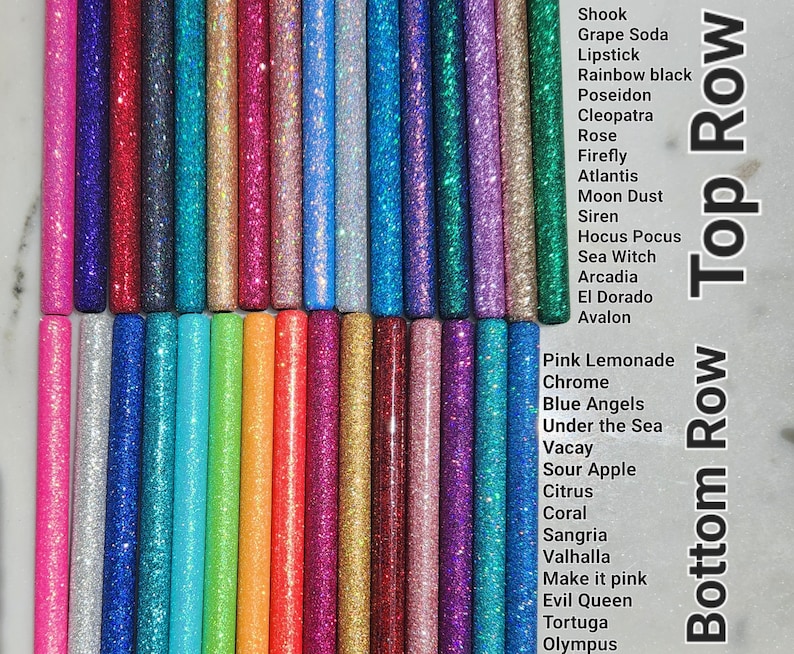 May include: A collection of glitter-filled glue sticks in various colors, arranged in two rows. The top row includes colors like Shook, Grape Soda, Lipstick, Rainbow Black, Poseidon, Cleopatra, Rose, Firefly, Atlantis, Moon Dust, Siren, Hocus Pocus, Sea Witch, Arcadia, El Dorado, and Avalon. The bottom row includes colors like Pink Lemonade, Chrome, Blue Angels, Under the Sea, Vacay, Sour Apple, Citrus, Coral, Sangria, Valhalla, Make it Pink, Evil Queen, Tortuga, and Olympus.