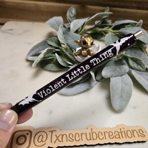 May include: A black and purple glitter pen with white bats and the text "Violent Little Thing" on it. The pen is held in a hand over a wooden sign with the text "@Txnscrubcreations" on it.