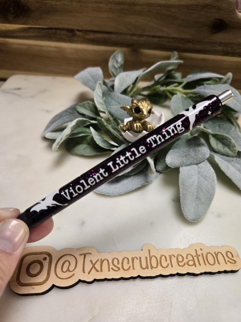 May include: A black and purple glitter pen with white bats and the text "Violent Little Thing" on it. The pen is held in a hand over a wooden sign with the text "@Txnscrubcreations" on it.