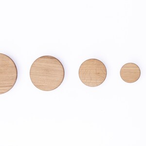 Puede incluir: Cinco ganchos de pared de madera de diferentes tama&ntilde;os, dispuestos en fila del m&aacute;s grande al m&aacute;s peque&ntilde;o. Los ganchos son circulares y de madera marr&oacute;n claro.