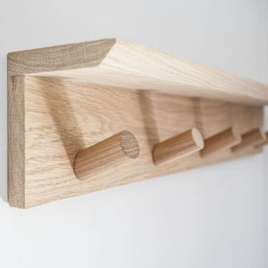 Könnte beinhalten: Ein helles Holzregal mit vier Holzhaken. Das Regal ist an einer weißen Wand montiert.