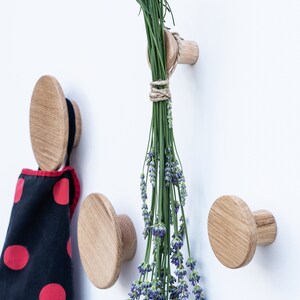 Puede incluir: Cuatro ganchos de pared de madera con un dise&ntilde;o redondo y plano. Un gancho tiene lavanda seca colgando de &eacute;l. Los ganchos est&aacute;n montados en una pared blanca.