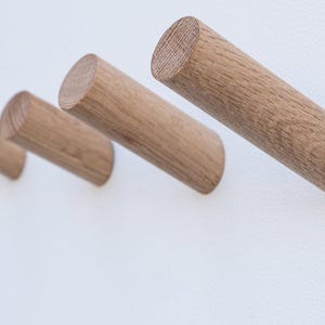 May include: Four wooden wall hooks with a natural wood grain finish are mounted on a white wall. The cylindrical hooks are angled slightly downward, providing a minimalist and modern design. The hooks are evenly spaced.