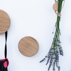 Puede incluir: Tres ganchos de pared de madera de diferentes tama&ntilde;os, con un delantal de lunares negros y rojos colgado del gancho m&aacute;s grande y un ramo de lavanda seca colgado del gancho m&aacute;s peque&ntilde;o.