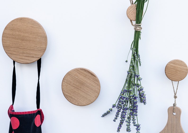 Puede incluir: Tres ganchos de pared de madera de diferentes tama&ntilde;os, con un delantal de lunares negros y rojos colgado del gancho m&aacute;s grande y un ramo de lavanda seca colgado del gancho m&aacute;s peque&ntilde;o.