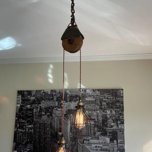 May include: Industrial-style pendant light fixture with two Edison-style bulbs encased in black metal cages. The fixture hangs from a pulley system with a wooden wheel and a dark metal chain, creating a vintage aesthetic. The background features a black and white cityscape.