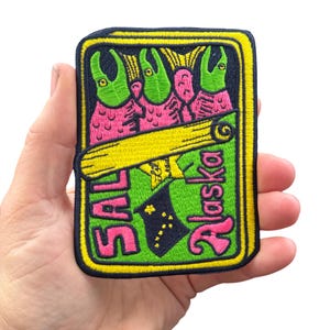 May include: Embroidered patch featuring a colorful design with the text "SAL Alaska". The patch has a black border with a yellow outline. The design includes green and pink figures, a yellow scroll, and the Alaska state flag.
