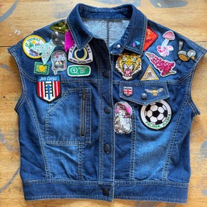 Puede incluir: Chaqueta vaquera sin mangas en un lavado azul medio, adornada con varios parches bordados. Los parches incluyen una cara de tigre, un balón de fútbol, una seta y un pájaro. La chaqueta tiene cremallera y botones.