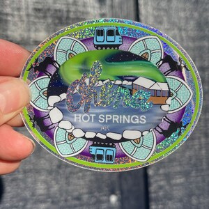 May include: A round, iridescent sticker with a green border. The sticker features a blue train, a snowy landscape, and the text "Chena Hot Springs AK". The background is a purple and blue mandala design.
