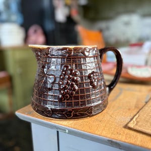 Könnte beinhalten: Ein brauner Keramik-Krug mit weißem Rand und einem Traubenmuster. Der Krug hat ein quadratisches Gittermuster und steht auf einem Holztisch.