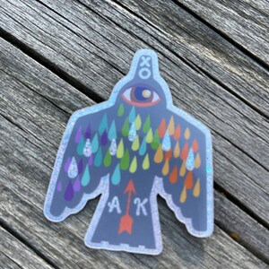 May include: A sticker with a bird design, featuring a large eye and colourful teardrops. The bird has outstretched wings and the letters "A K" with an arrow. The sticker has a holographic, glittery border and the word "XO" at the top.