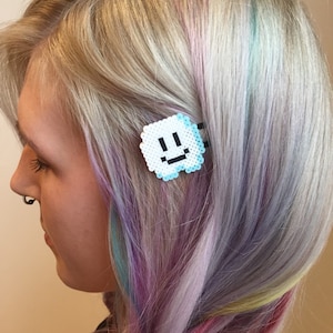 Op de afbeelding: Een witte en blauwe gepixelde smiley haarspeld. De speld zit in het haar van een vrouw met paarse, blauwe en gele strepen.