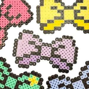 Op de afbeelding: Een verzameling kleurrijke pixel art strikjes in verschillende tinten roze, geel, groen en blauw. Elk strikje is omlijnd in zwart en gemaakt van kleine, gesmolten plastic kralen, wat een retro, handgemaakte uitstraling creëert.