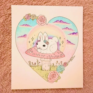 以下が含まれることがあります： ピンクのリボンをつけた白いウサギが、白い斑点のあるピンクのキノコの上に座っています。キノコは、虹と雲のあるハート型のフレームの中にあります。背景は薄いピンク色で、花が咲いています。