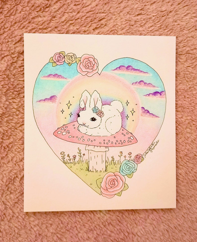 以下が含まれることがあります： ピンクのリボンをつけた白いウサギが、白い斑点のあるピンクのキノコの上に座っています。キノコは、虹と雲のあるハート型のフレームの中にあります。背景は薄いピンク色で、花が咲いています。
