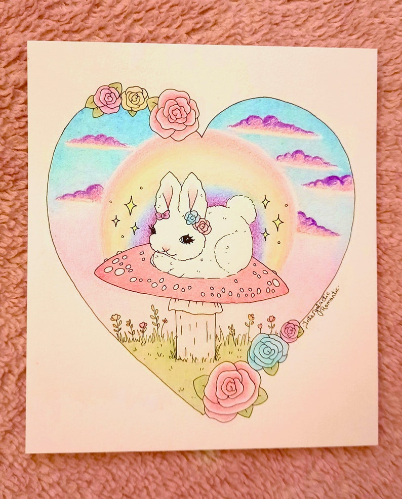 以下が含まれることがあります： ピンクの花を頭につけた白いウサギが、緑の茎のついた赤と白のキノコの上に座っています。キノコは緑の草とピンクと青の花に囲まれています。背景は虹と雲のあるハート型です。