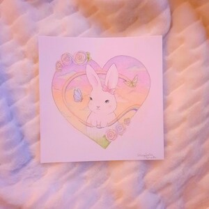 Puede incluir: Un conejo blanco con un lazo rosa se sienta dentro de un marco en forma de coraz&oacute;n. El marco est&aacute; lleno de un arco&iacute;ris, nubes y mariposas. El fondo es de color rosa suave.