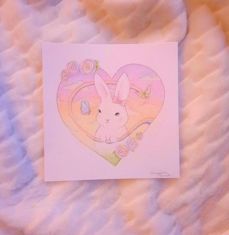 Puede incluir: Un conejo blanco con un lazo rosa se sienta dentro de un marco en forma de coraz&oacute;n. El marco est&aacute; lleno de un arco&iacute;ris, nubes y mariposas. El fondo es de color rosa suave.