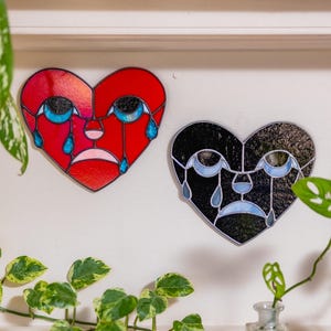 May include: Two heart-shaped stained glass art pieces. One is red with blue teardrops and a sad face, the other is black with blue teardrops and a sad face. The art pieces are mounted on a white wall.
