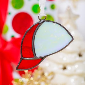 May include: A stained glass baseball cap ornament with a red brim and a white and opalescent crown. The ornament is trimmed in black and hangs from a white ribbon. A festive holiday decoration.