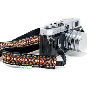 May include: A silver Canon camera with a black leather body and a black and orange patterned camera strap. The strap has the text "ELLUM BAG WORKS" on it.