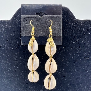 May include: A pair of gold-toned earrings with three white cowrie shells on each earring. The shells are strung on wire and the earrings have a simple hook closure.