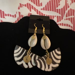 May include: A pair of earrings with a black and white zebra print design. The earrings are made of wood and have a gold wire accent. The earrings are hanging from a gold hook.