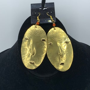 May include: A pair of gold oval-shaped earrings with a textured surface and small gold accents. The earrings are hanging from a black display card.