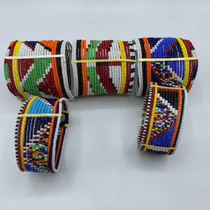 May include: A collection of five wide cuff bracelets crafted from small, multi-colored glass beads. The bracelets feature intricate geometric patterns in vibrant shades of blue, red, green, orange, yellow, white, and black, arranged in repeating triangular and striped designs.