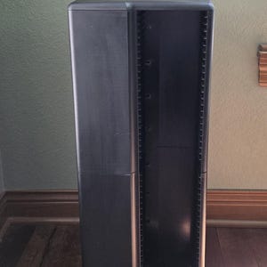 May include: A tall, black, rectangular media storage tower featuring a vertical column of individual slots designed for organizing compact discs or similar media cases. The unit has a smooth, matte finish and a sturdy, upright, freestanding structure.