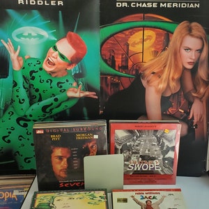 Op de afbeelding: Een verzameling film-dvd's en VHS-banden met acteurs Jim Carrey, Nicole Kidman, Brad Pitt, Morgan Freeman en Robert Downey Jr. Titels zijn onder meer "Seven", "Chinatown" en "Jack". De achtergrond bevat filmposters.