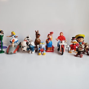 Può includere: Una collezione di piccole e colorate statuette di personaggi dei cartoni animati. Le statuette includono personaggi Disney, Hanna-Barbera e altre serie animate. Le statuette sono di plastica e sono su una superficie bianca.